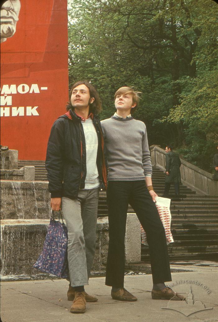 Два хлопці на тлі радянського агітаційного стенду 2